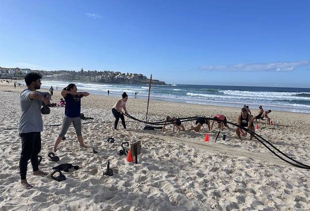 Group of people doing a high-intensity beach fitness workout with battle ropes and kettlebells at Bondi Beach.
