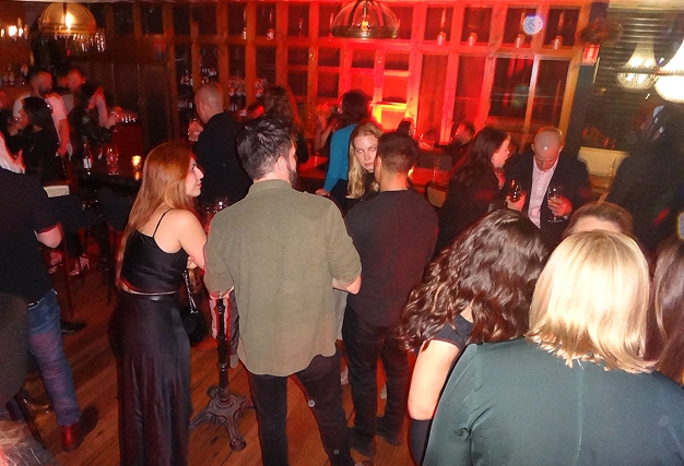 Social singles event atmosphere in a dimly lit bar with red ambient lighting, showing attendees mingling and drinking.