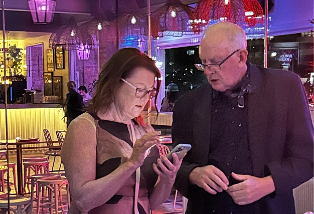 Man and woman (older demographic) at a bar, looking at a smartphone, likely checking in or confirming event details.