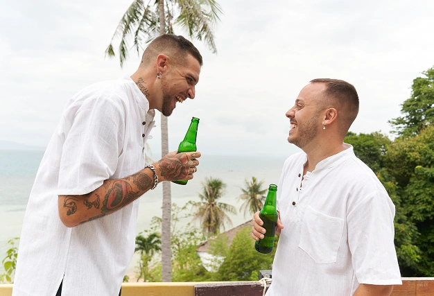 Two smiling men in white shirts toasting with bottled beer on a balcony with a tropical beach and ocean view, symbolizing casual fun.