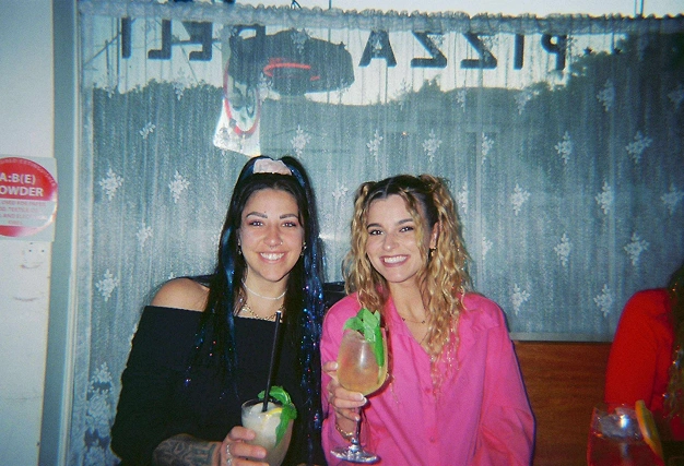Two happy women holding cocktails and smiling at the camera in a warmly lit bar, representing a casual singles night.