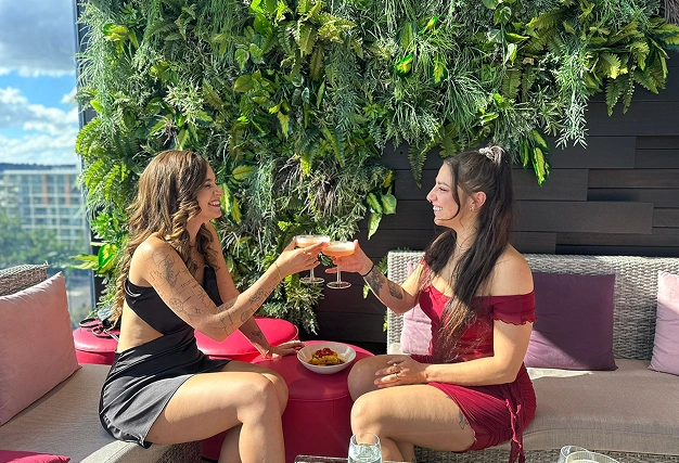 Two women toasting cocktails on a sunny rooftop bar with a vibrant vertical garden background, symbolising a chic singles meet-up.