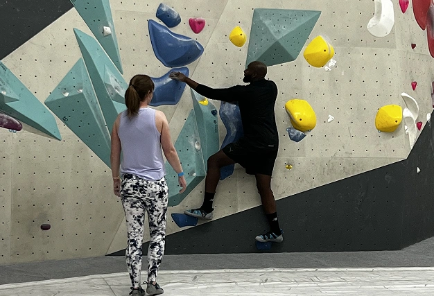 A man in black athletic wear climbs a grey bouldering wall while a woman in patterned leggings observes from below.