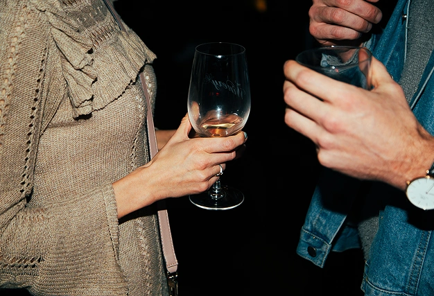 Close-up of a woman in a beige knit sweater holding a wine glass and a man in a denim jacket holding a drink.