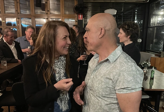 A woman with a leopard print scarf laughs while talking face-to-face with a man in a light blue floral shirt at a busy venue.