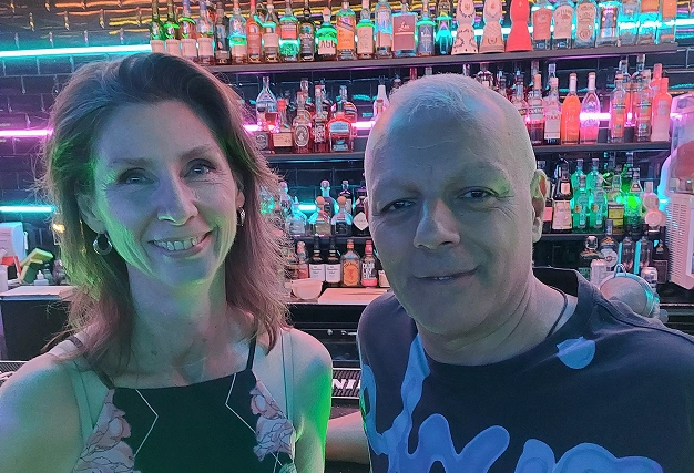 A man and a woman smiling for a portrait in front of a bar display shelf lit with vibrant blue and pink neon lights.