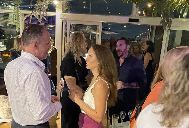 Guests socializing at a rooftop bar with glass walls, greenery hanging from the ceiling, and city lights visible outside.