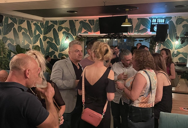 A crowded social event in a lounge with distinctive green banana leaf wallpaper and warm interior lighting.