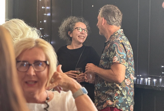 A woman with curly grey hair and glasses smiles warmly while talking to a man in a dark floral patterned shirt.