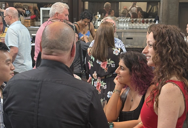 A woman in a red dress and a woman with curly hair leaning on her hand chat at a bar while others stand nearby.