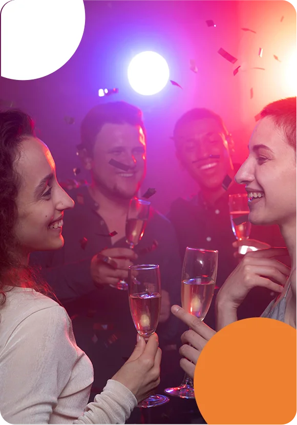 A group of diverse, smiling young adults celebrating at a party with purple and red stage lighting. They are holding champagne flutes for a toast as confetti falls around them. The image features a modern graphic style with large white and orange circular overlays in the corners.